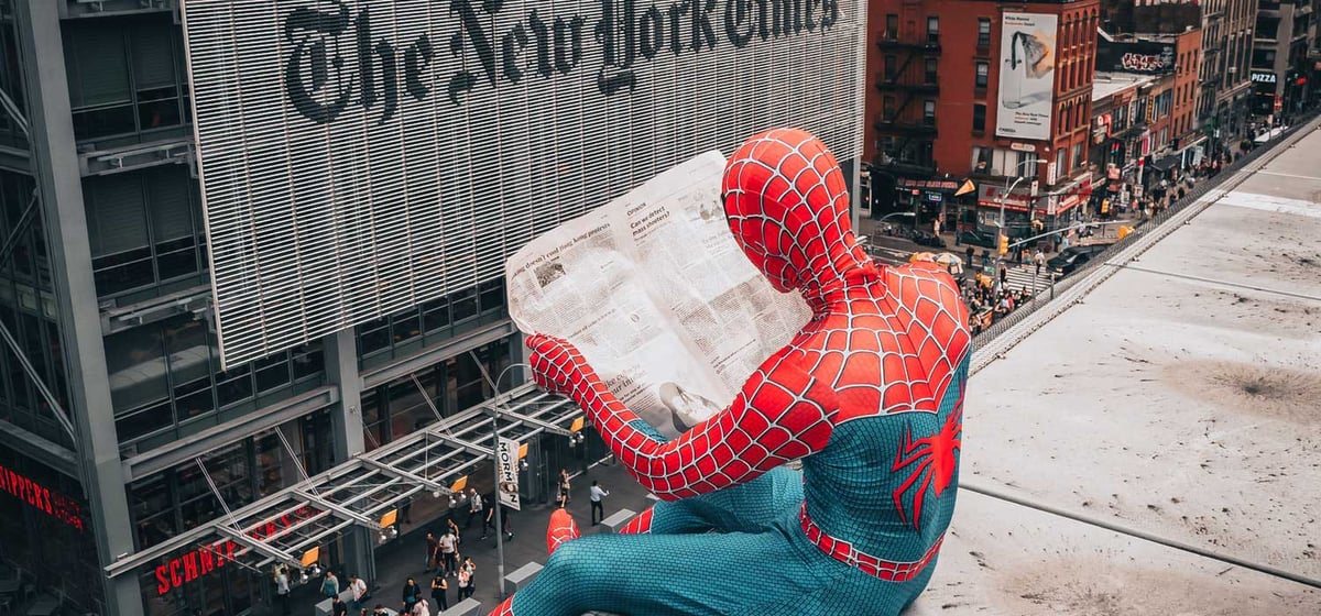 Spider-Man sits on the ledge of a rooftop while he reads a newspaper.