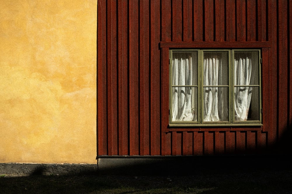 Falurött trähus med ett fönster med vita gardiner bredvid en solbelyst ockragul putsad vägg, Stockholm