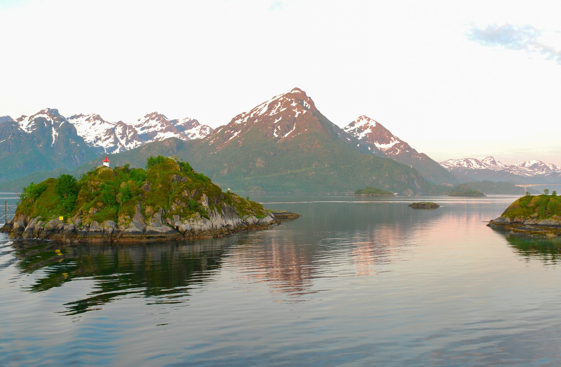 Green islet with a small lighthouse, still water mirroring snowy peaks, Lofoten